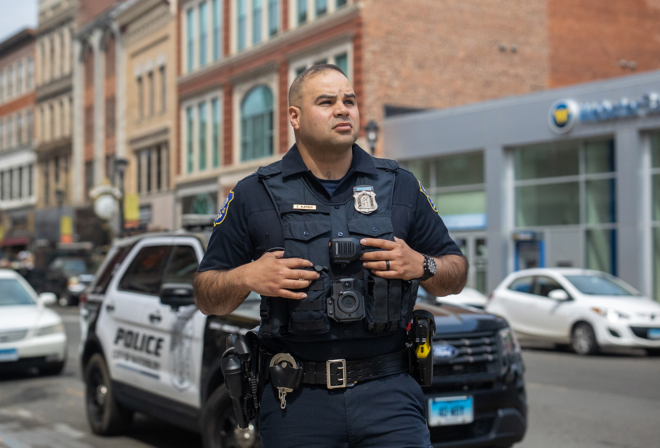 A Waterbury police officer standing on a city street in front of a patrol vehicle