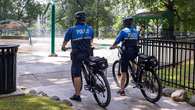 Two Waterbury police officers on bicycles stopped in a park near a splash pad